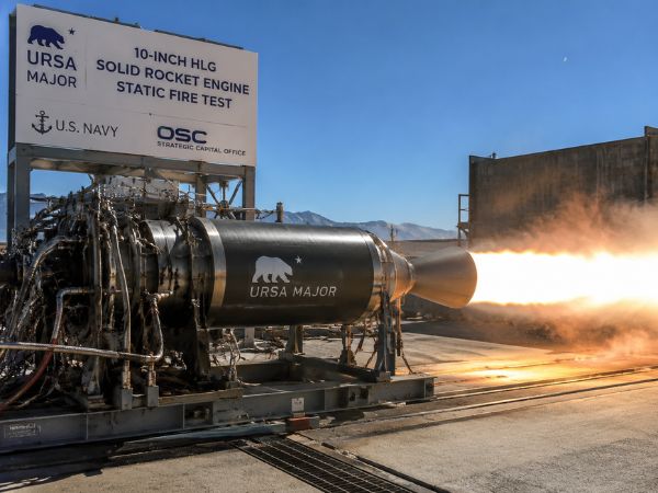 A metal 3D printed solid rocket engine prototype undergoing a static fire test at an outdoor aerospace testing facility, with bright exhaust flames, industrial equipment, and Ursa Major branding visible on the engine and test structure.