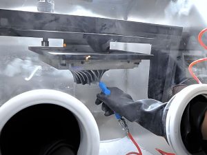 Operator removing residual powder from a metal 3D printed part inside a sealed glovebox using compressed gas.