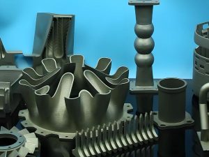 Array of complex metal 3D printed industrial components including a multi-flanged aero-engine nozzle, turbine impeller, and thin-walled structural parts.